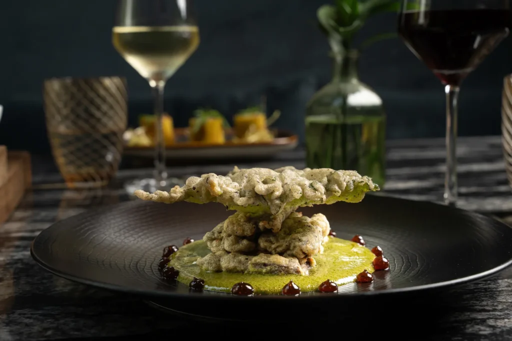 A black plate with a gourmet dish featuring tempura-fried vegetables on green sauce, garnished with brown sauce dots; perfect for social gatherings, with wine glasses and blurred background decor adding to the ambiance.